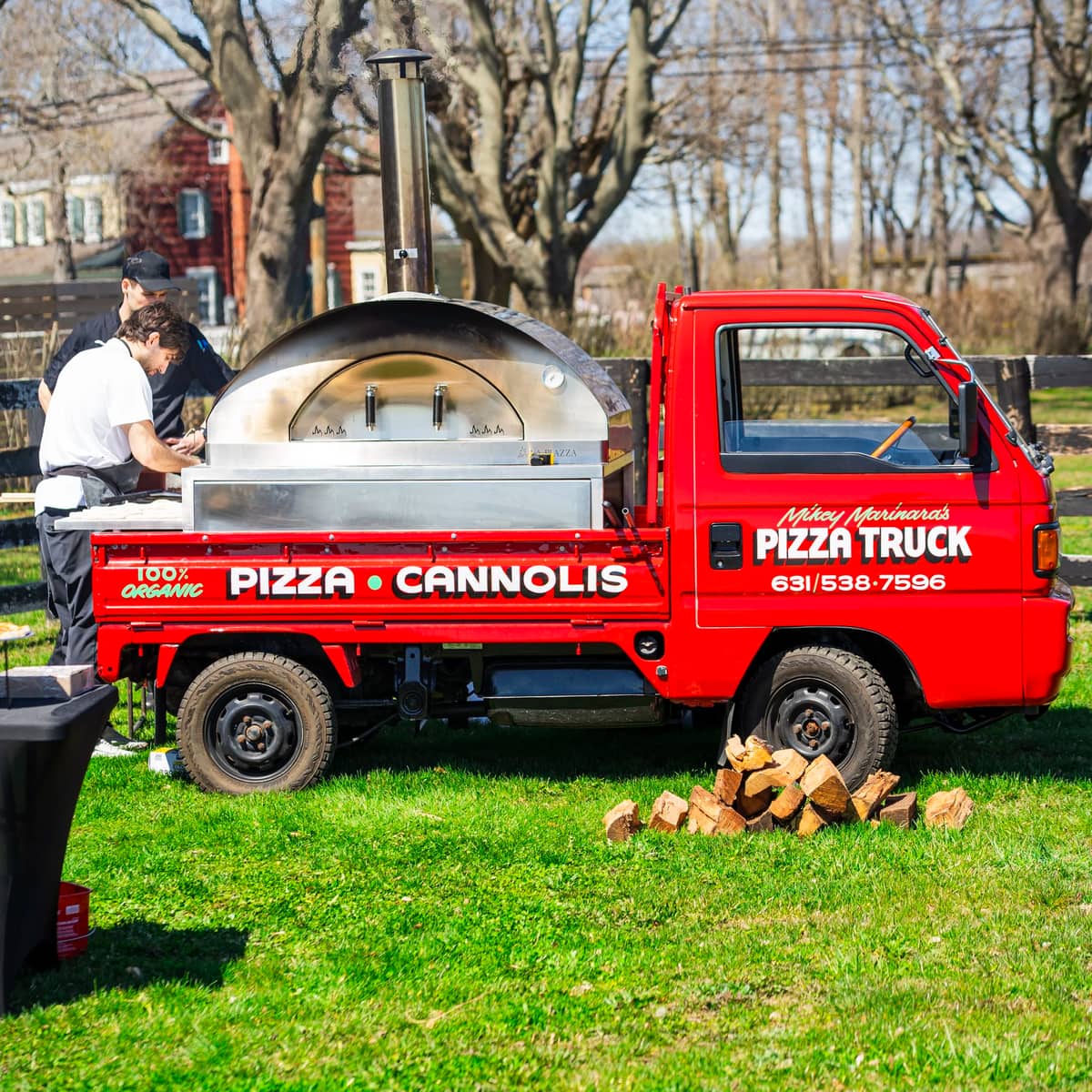 The red pizza truck parked on a back lawn with its wood-fired oven lit and split firewood stacked beside the wheel