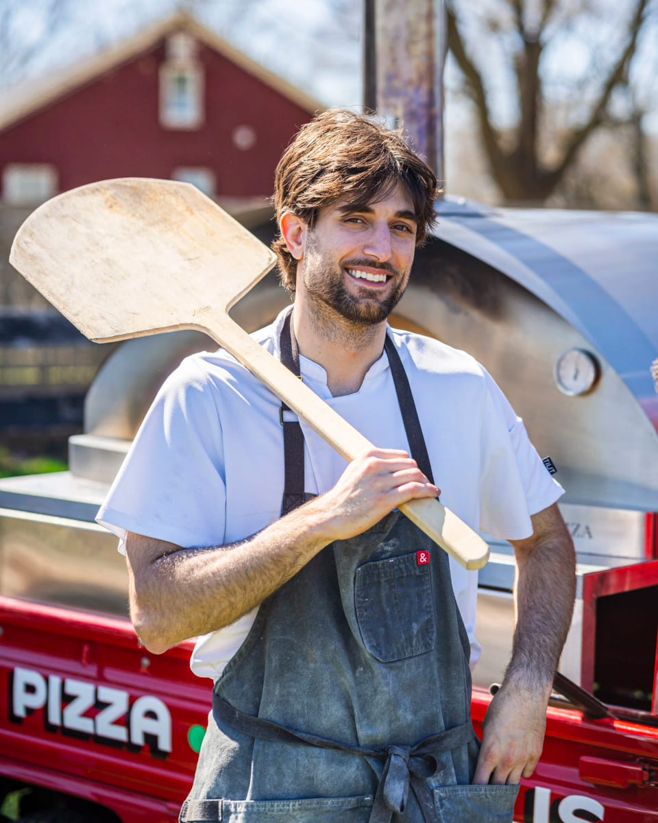 Mikey holding a wooden pizza peel beside a red barn