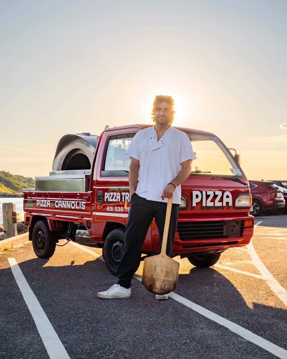 Mikey with a pizza peel in front of the truck at a Long Island harbor at sunset