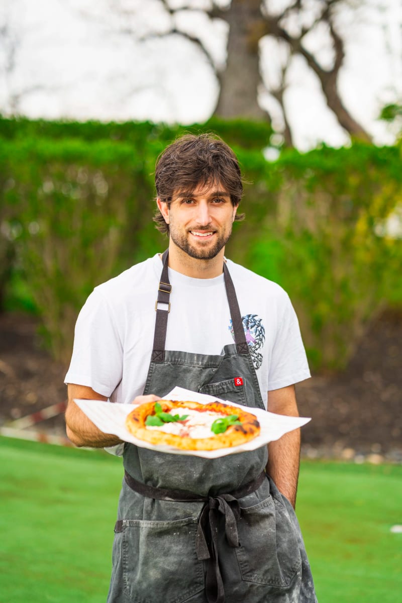 Mikey holding a freshly fired pizza on paper, in his apron